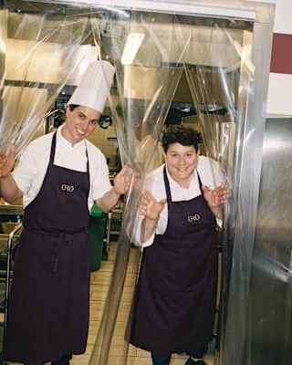 Wearing black aprons, Head Chef Vania Ghedini and a male chef in a white toque peer through kitchen partition curtains.