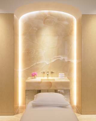 The strip lighting around an alcove illuminates a marble sink with a bud vase and flannels, seen over a bed with pillow.