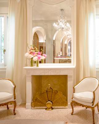 A chandelier reflects in a mirror above a fireplace alcove flanked by rococo white and gold armchairs, in a bright spa room.