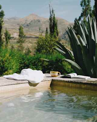A white towel, mug and book rest on the corner edges of a pool in an eye-level view from the water, capturing hills beyond.