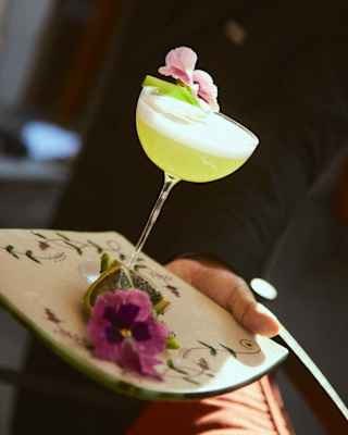 A waiter holds a ceramic tray with a lime-green pisco sour cocktail, garnished with a pink pansy, in an angled close-up.