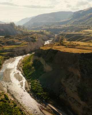 Sunlight glistens on the Colca river as it snakes between the green terraces and rock cliffs of the canyon, seen from above.