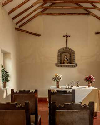View over four wood and leather seats to a small chapel altar with a white cloth and flower vases either side of two goblets.