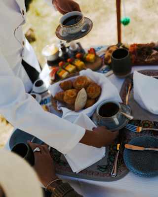 A person in a white shirt serves coffee and pastries to someone wearing a hat at an outdoor table set with cups, plates, and assorted breakfast foods.