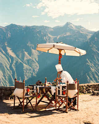 A dining table with chairs and a large parasol is set up outdoors on a mountaintop, overlooking scenic mountains under a partly cloudy sky. The table is decorated for a meal, creating a picturesque setting.