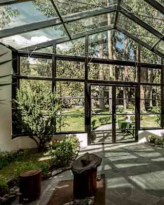 A sunroom with glass walls and ceiling, filled with potted plants and greenery along the edges. Outside, a lush garden with trees is visible. The floor is stone, and there are rustic wooden stools and a bench.