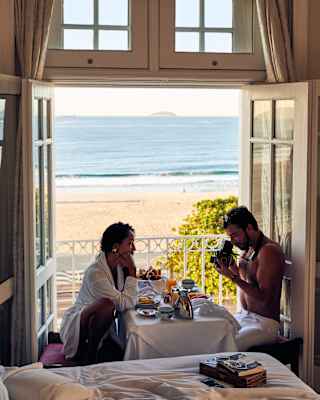 A woman sitting at the breakfast table facing her partner while he takes photographs of her with a vintage camera