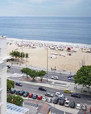 Calm blue sea laps the bustling Copacabana Beach. People stroll the sidewalk. Its pattern is repeated on the hotel's forecourt