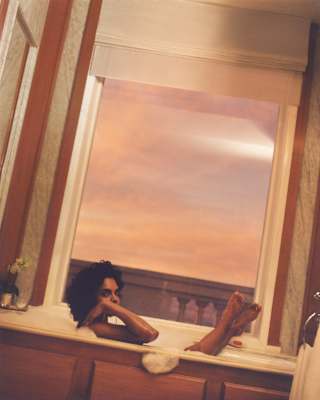 A woman with dark curly hair soaks in a foam bath with her feet up on the sill of the bathroom window, framing the sunset.