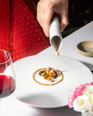 A waiter pours jus on a pretty mushroom dish with edible flower garnish for a diner at a table with red wine, seen close-up.