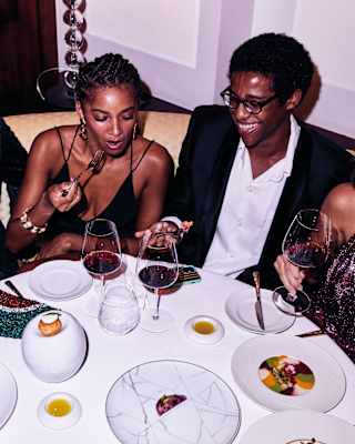 Five young people in party dress sit at a round table laughing and drinking red wine while sharing plates of dessert