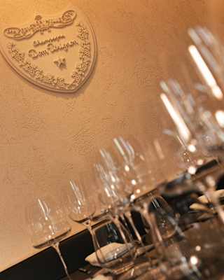 Angled image of a Dom Perignon plaque in the brand's trademark shield-shape on the wall above a table of empty glasses.