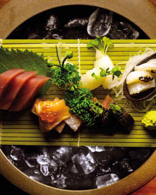 Assorted sashimi pieces and garnishes arranged on a bamboo mat over a bowl of ice, with wasabi and seaweed, set on a wooden table.