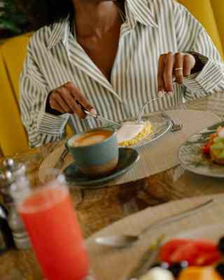 Angled eye-level view over a breakfast to a woman in a green-striped shirt, seen from the neck down, enjoying scrambled eggs.
