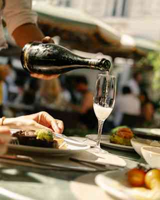 A person pours sparkling wine into a flute glass at an outdoor restaurant table set with crockery, with blurred people and umbrellas in the background.