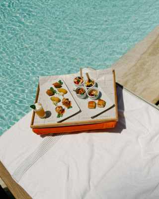 A tray with a drink and two plates of assorted appetisers, with fruit pots and pastry squares, rests on a poolside lounger.