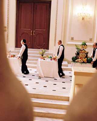 Three uniformed staff members push room service tables with drinks and fruit across a hallway, seen through banisters above.