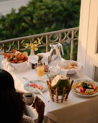 A table with fruit, pastries and a coconut with straw in a bamboo stand, seen over the shoulder of a woman dinking coffee.