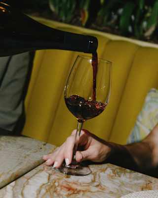 A hand holding a wine glass as red wine is being poured into it, with a yellow cushioned banquette and a marble table in the background.