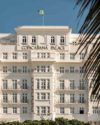 The white facade of Copacabana Palace hotel with many windows, a Brazilian flag on the roof, and a palm frond partially visible in the foreground against a clear blue sky.