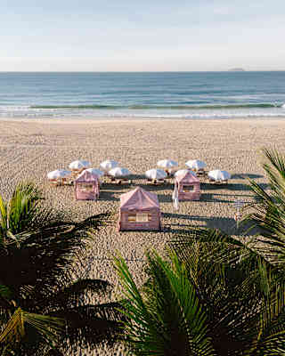 Three red-striped cabanas and ten pairs of loungers shaded by white parasols face the sea in a view of the beach over palms.
