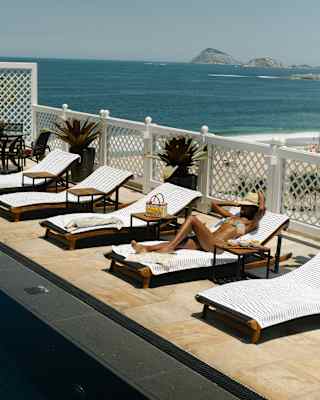 A tanned woman in a bikini and straw sunhat reclines on a towel-covered poolside lounger with views beyond of the beach.
