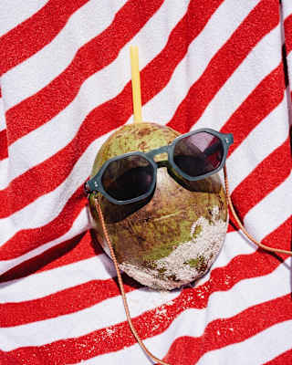 Comical photo of a sandy coconut on a red and white-striped towel sporting sunglasses on a chain, with a straw in its head.