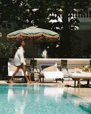 A guest, blurred by motion, walks past reclining sun-loungers at the edge of the pool, seen at eye-level across the water.