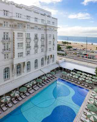 High-angle view of a large hotel swimming pool with loungers and parasols, situated next to a historic white building and a beach.