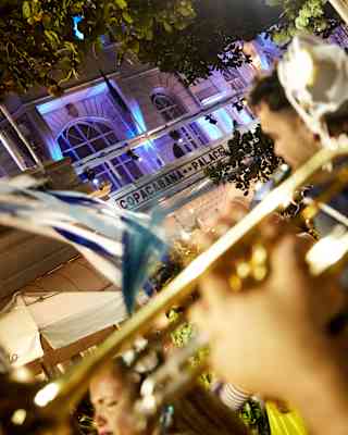 Angled image of a busy street outside Copacabana Palace at night, illuminated by blue spotlights, seen over a brass trombone.