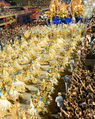 Looking down on a carnival parade of dancers in tufty yellow and blue costumes in front of a huge float, watched by crowds.