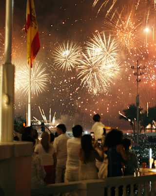A group of men, women and children stand on the terrace watching firework explosions in a night sky, seen from behind.