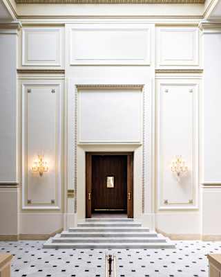A monochrome tiled floor and five steps lead to theatre's doors, flanked by lights, set in a tall white, wood-panelled wall.