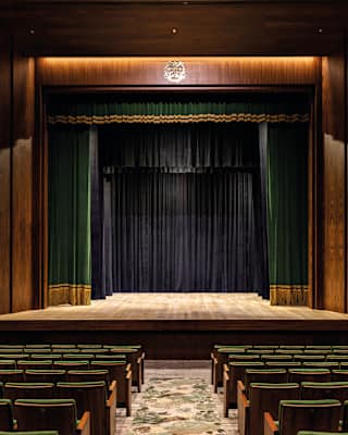 Green velvet curtains, edged in gold trim, are open on wood panelled stage, faced by rows of green velvet theatre seating