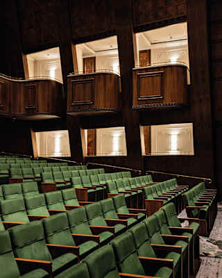 Neat rows of green velvet fold-down theatre chairs climb the gentle incline of the floor, watched by wood panelled boxes