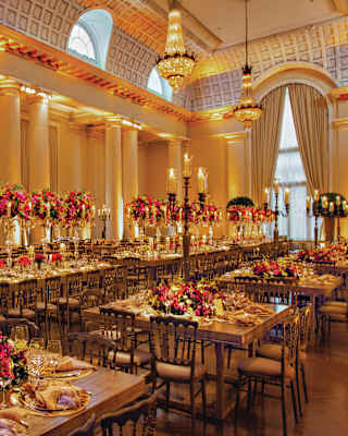 Grand banqueting hall with high ceilings, large windows, chandeliers, and long tables elegantly laid with flowers, candles, and place settings, ready for a formal event or celebration.