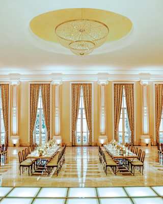 Four long celebration tables await guests in a room with five tall glazed doors with bronze drapes, and honey marble floors.