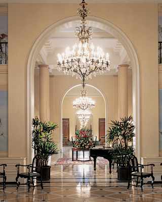 A row of blazing Bohemian chandeliers, seen between the high wall-arches of a lobby adorned with murals and antique chairs.