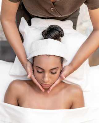 A spa therapist in a brown uniform cups the face of a guest wearing a head towel, during a facial treatment, seen from above.