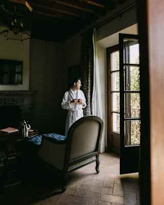Daylight spills through glass doors into a low-lit suite, illuminating a man in a robe, who gazes out, holding a coffee cup.