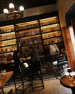 In a wood-toned room, a couple sits at a bar as a barman stands aside in front of backlit bottle shelves, seen on an angle.