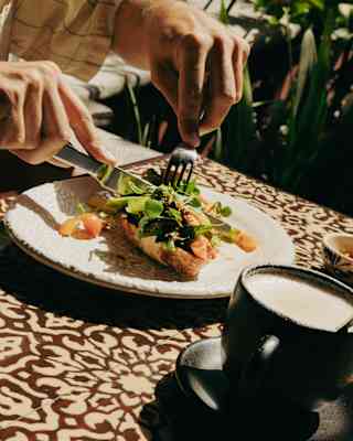 Angled image of a man's hands holding a knife and fork as he cuts into a tosta de aguacate, served with coffee at Del Parque.