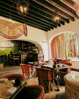View across a dining room with warm hues and wooden beams, with one wall arches leading to a bar and another to gardens.