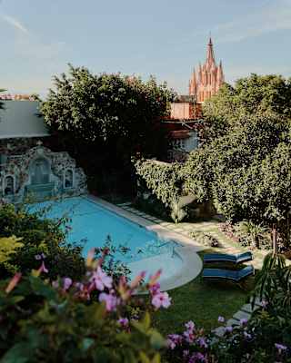 Sun illuminates the sugar-pink spires of Parrocchia San Michele Arcangelo in a high view over the shaded gardens and pool.