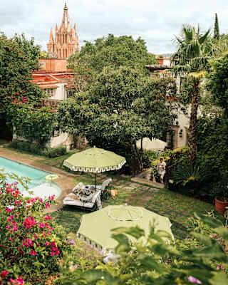 A man reads on a poolside sun-lounger, in a high-angle view that captures the verdant gardens and pink church spires beyond.