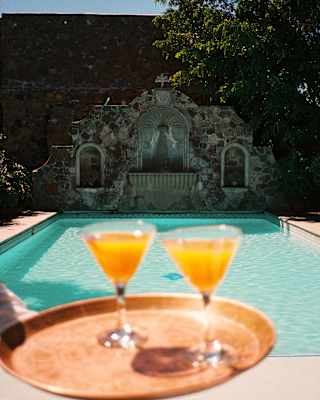 A waiter carries two orange drinks on a copper tray, seen close-up in soft-focus, with the defined sparkling pool behind.