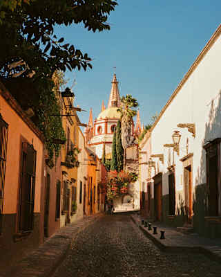 Sun illuminates buildings, domes and the iconic pink church spires, viewed through the shady cobbled street of Calle Aldama.