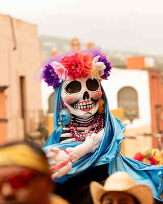 A skull-faced puppet wearing a red, pink and purple floral headband and blue cloak is held above crowds in a street parade.