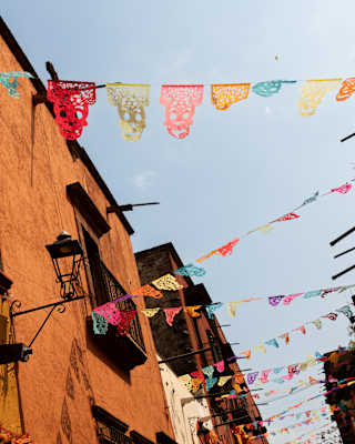 Strings of bright papel picado zigzag between the orange-rendered buildings of a San Miguel street, seen from below.