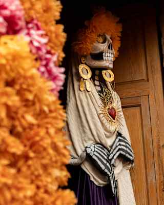 A Dia de Muertos skull-head figure stands in a doorway in large earrings and a necklace, part-obscured by a marigold garland.
